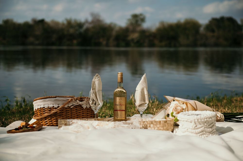 Romantic lakeside picnic setting with wine bottle, glasses and woven baskets on white cloth at dusk.