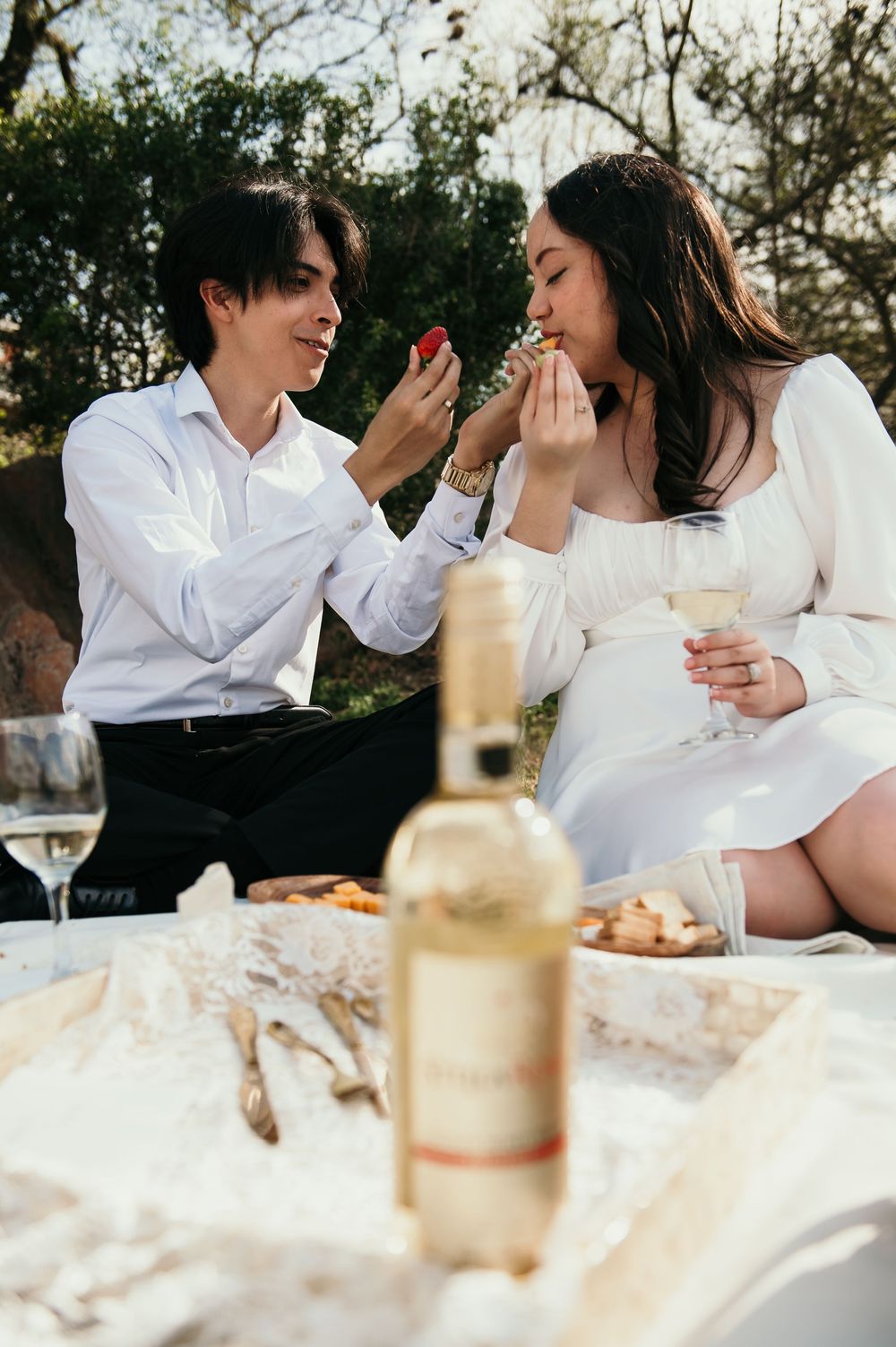 A romantic outdoor picnic scene with wine and sharing food on a blanket at sunset.