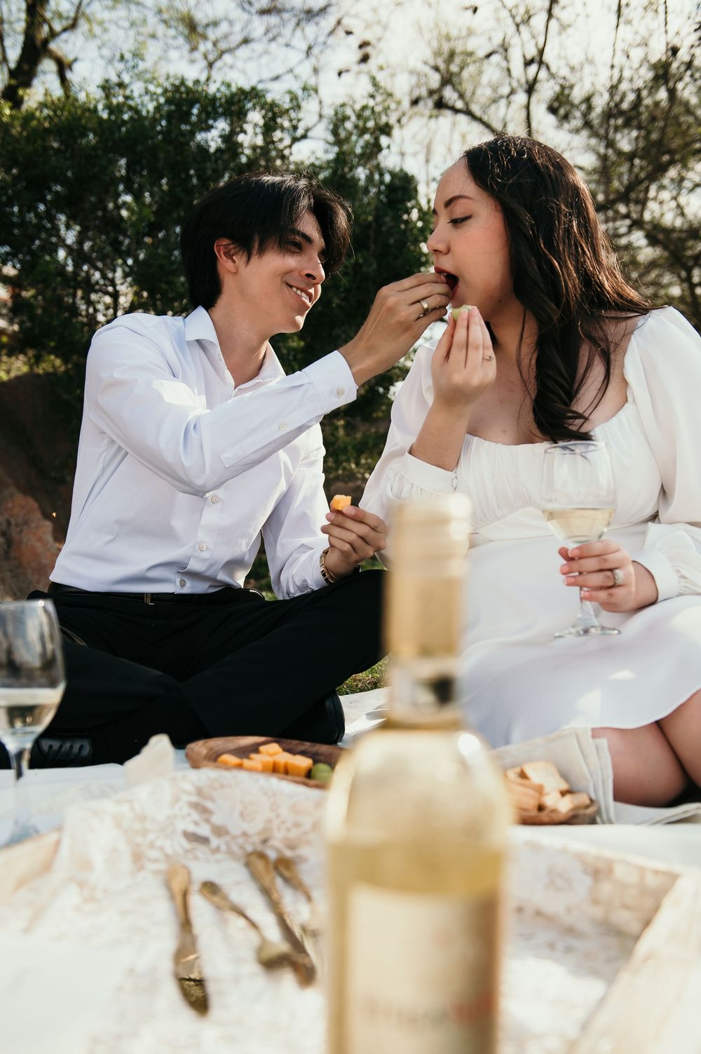 Couple in white attire sharing a romantic picnic moment while feeding each other outdoors at sunset.