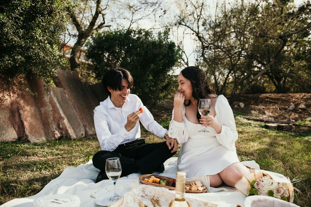 A romantic outdoor picnic scene with a white blanket, food and refreshments laid out under trees.