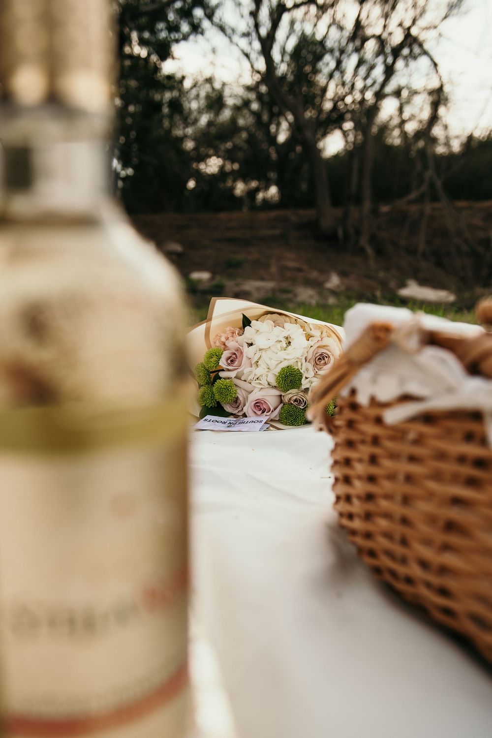 Close-up of wine bottle and gourmet sandwich on white tablecloth with picnic basket and trees in background at dusk.