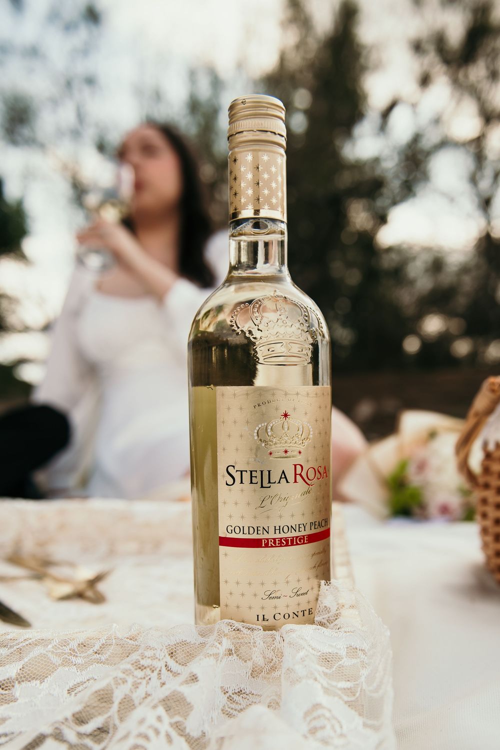 A bottle of Stella Rosa Golden Honey Peach wine sits on a lace tablecloth during an outdoor gathering.