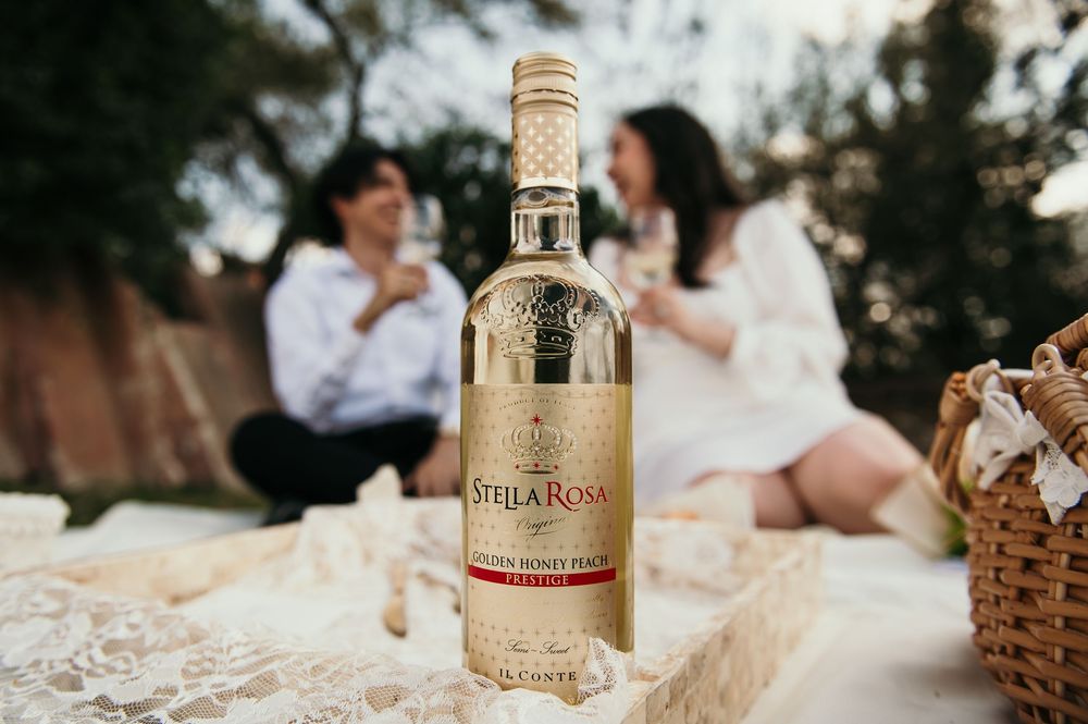A bottle of rose wine on a picnic blanket with a wicker basket and couple sitting in the background.