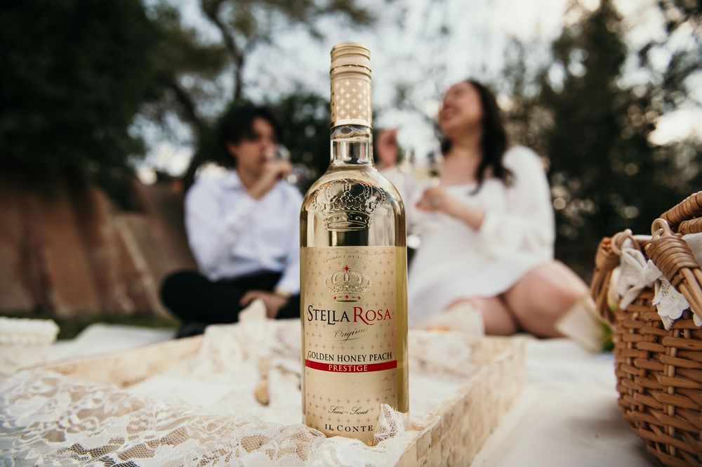 A bottle of Stella Rosa wine sits in foreground with a picnic scene and blurred figures in the background.