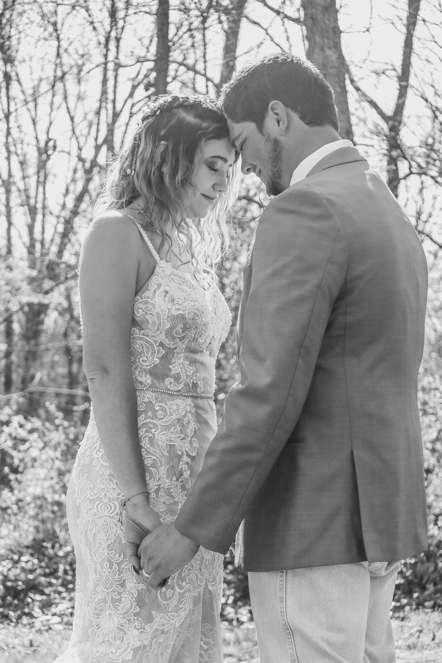 A romantic black and white photograph shows a couple in wedding attire embracing outdoors in a wooded setting.