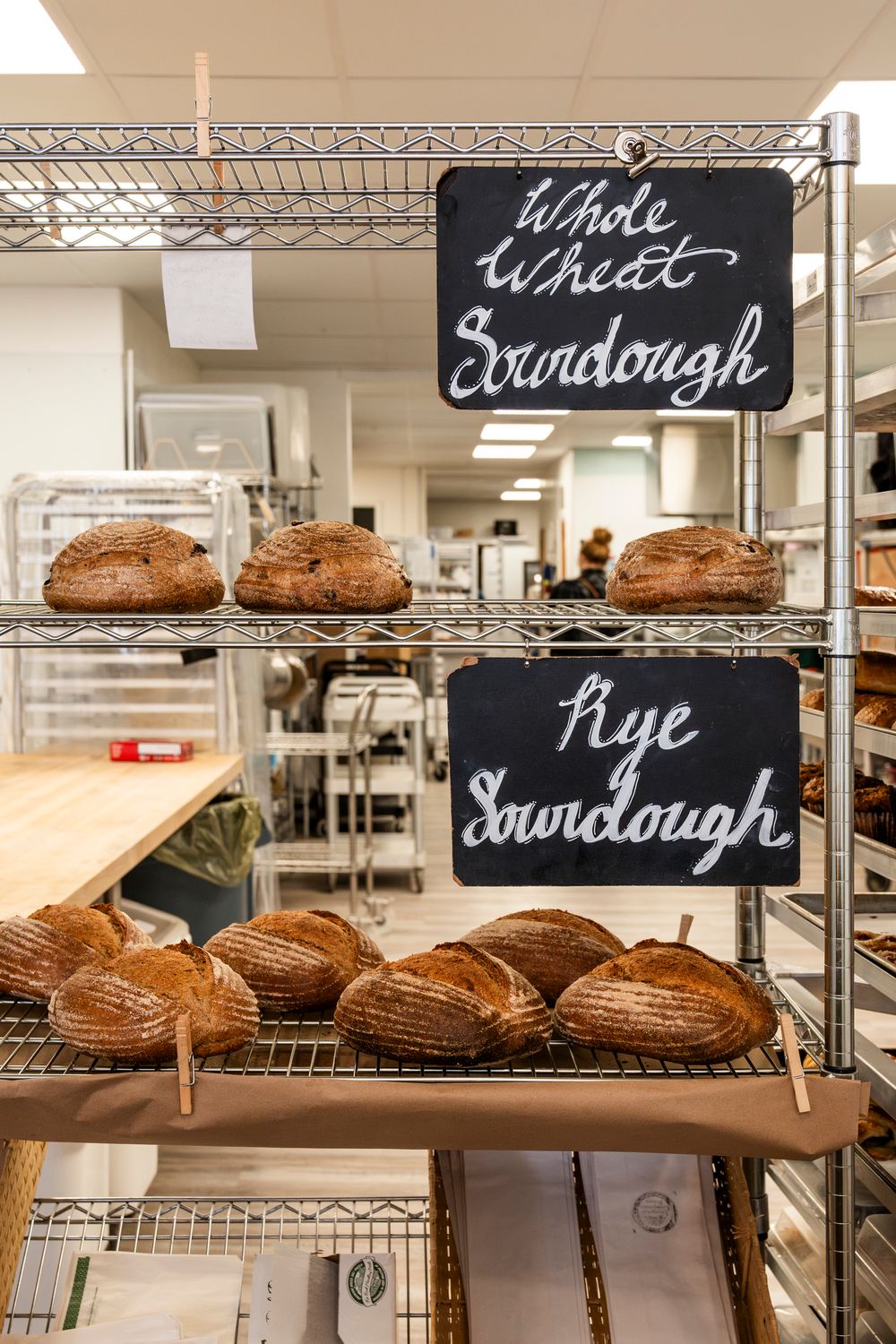 Artisanal sourdough and rye bread loaves displayed on wire racks with handwritten signs.