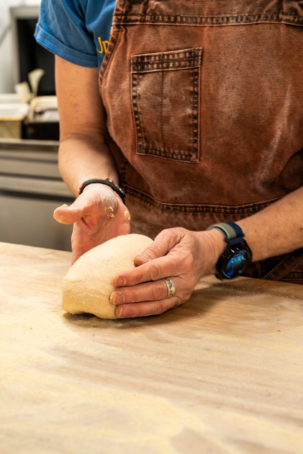 Hands in brown apron kneading raw bread dough on a wooden surface.