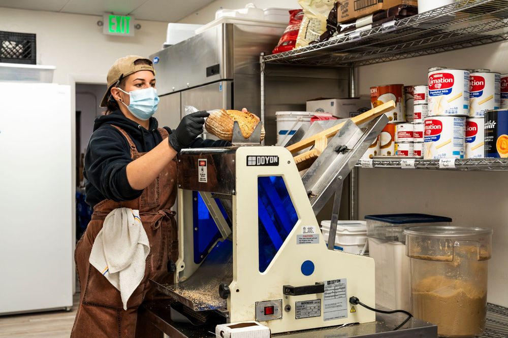Baker in protective mask operating industrial baking equipment in commercial kitchen.