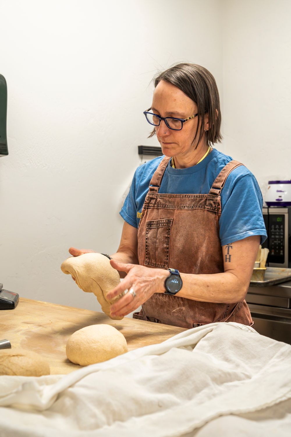 Baker in brown overalls shaping raw bread dough on a floured workspace.