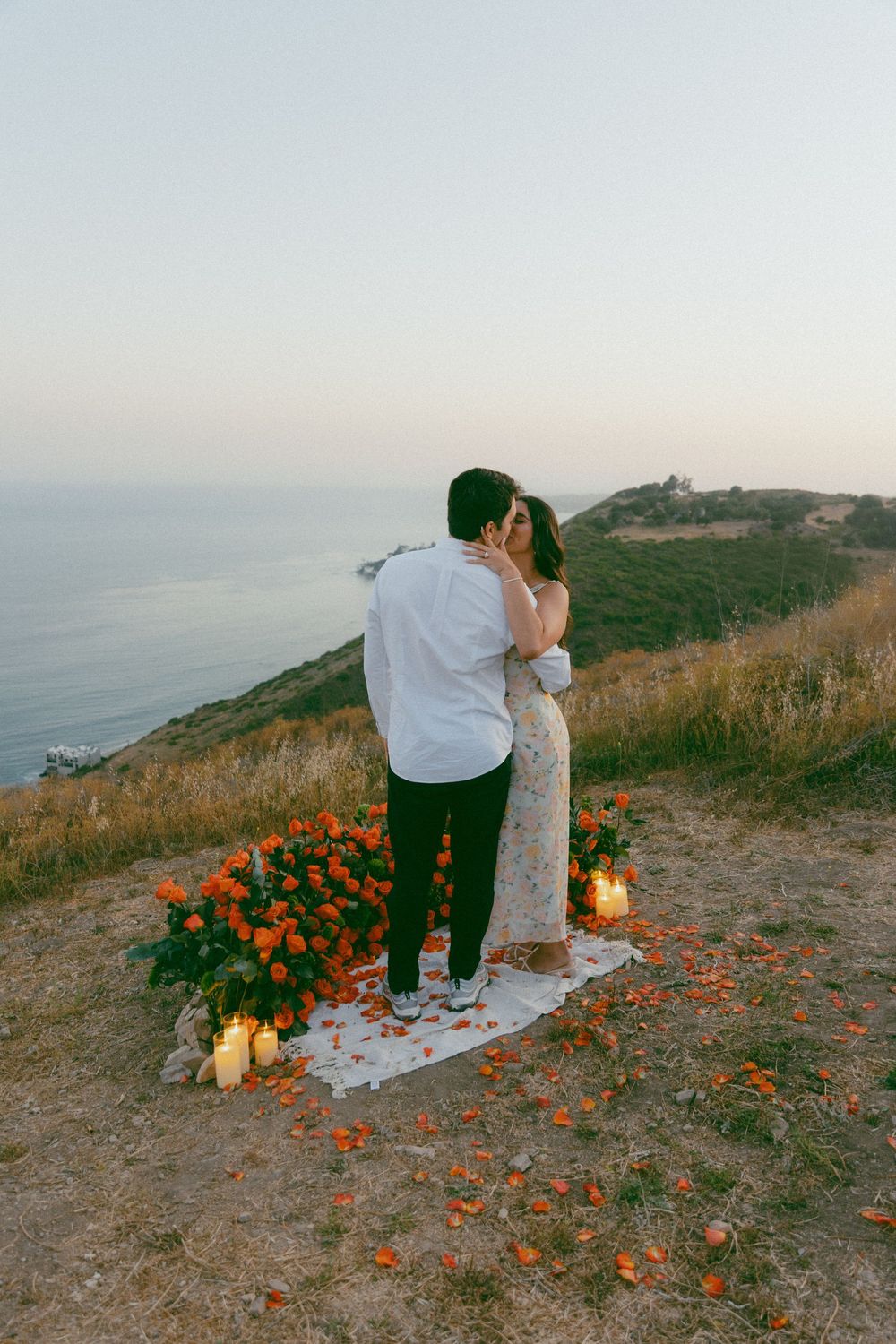 A romantic proposal setup on a coastal cliff at sunset with candles and orange flowers lining a white carpet path.