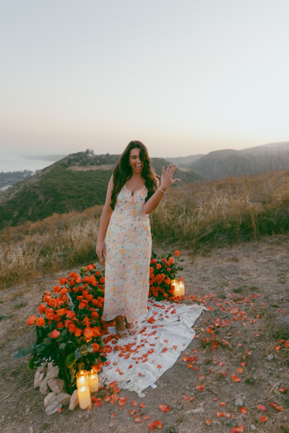 A scenic outdoor photoshoot with rose petals scattered on a blanket against a mountain backdrop at sunset with glowing candles.