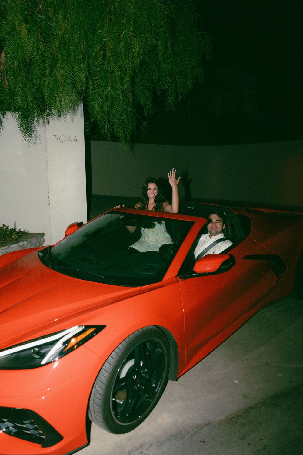 A bright red Chevrolet Corvette parked at night with two people waving from inside the sports car.