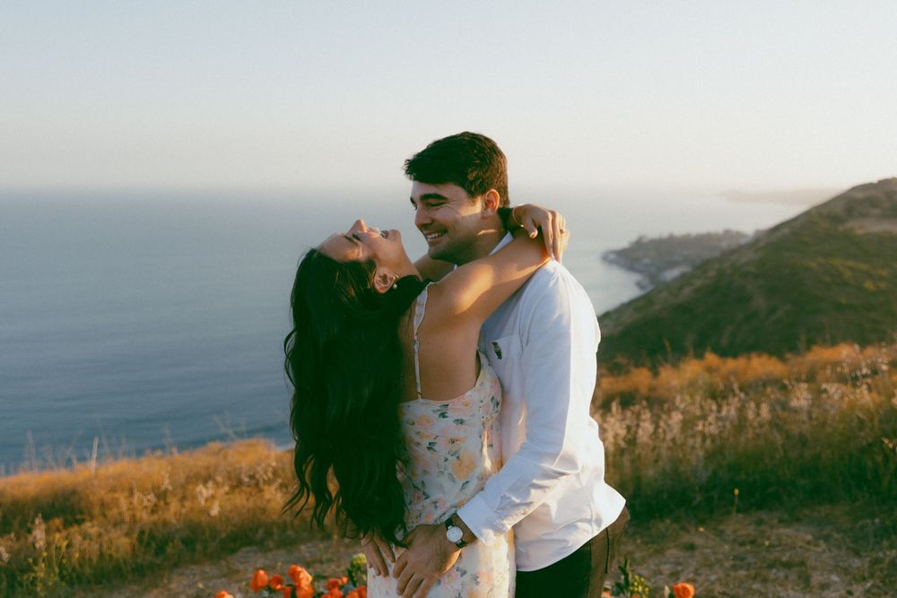 Couple embracing on a scenic coastal cliff during golden hour with ocean views in the background.