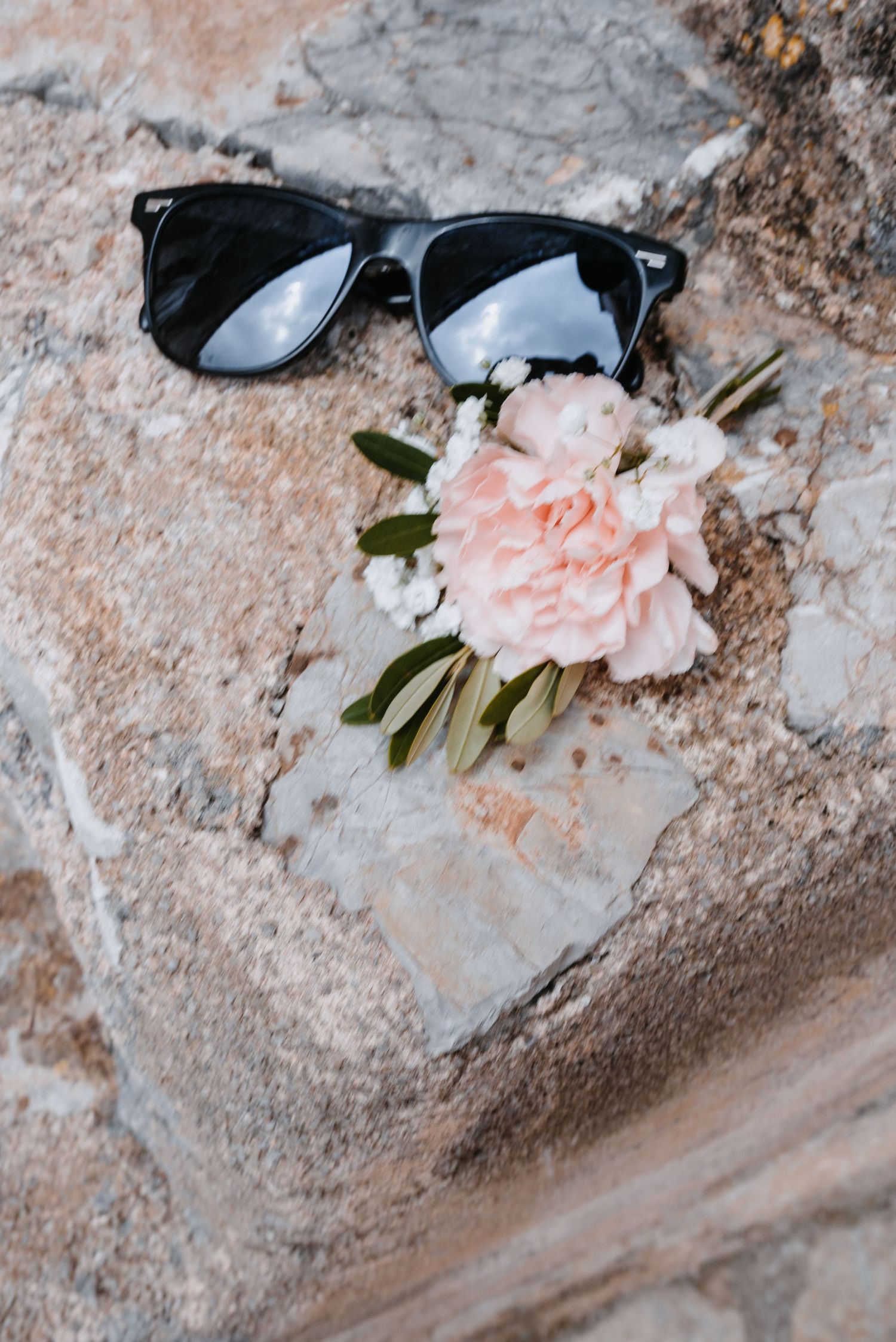 Detailbild mit schwarzer Sonnenbrille des Bräutigams und einem zarten, rosa Nelkenblüten-Anstecker auf einer strukturierten Steinoberfläche.