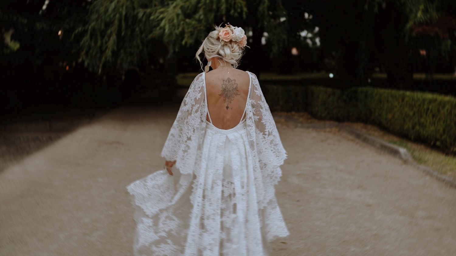 Elegant wedding dress with open back design and flowing cape sleeves photographed from behind on a garden path.