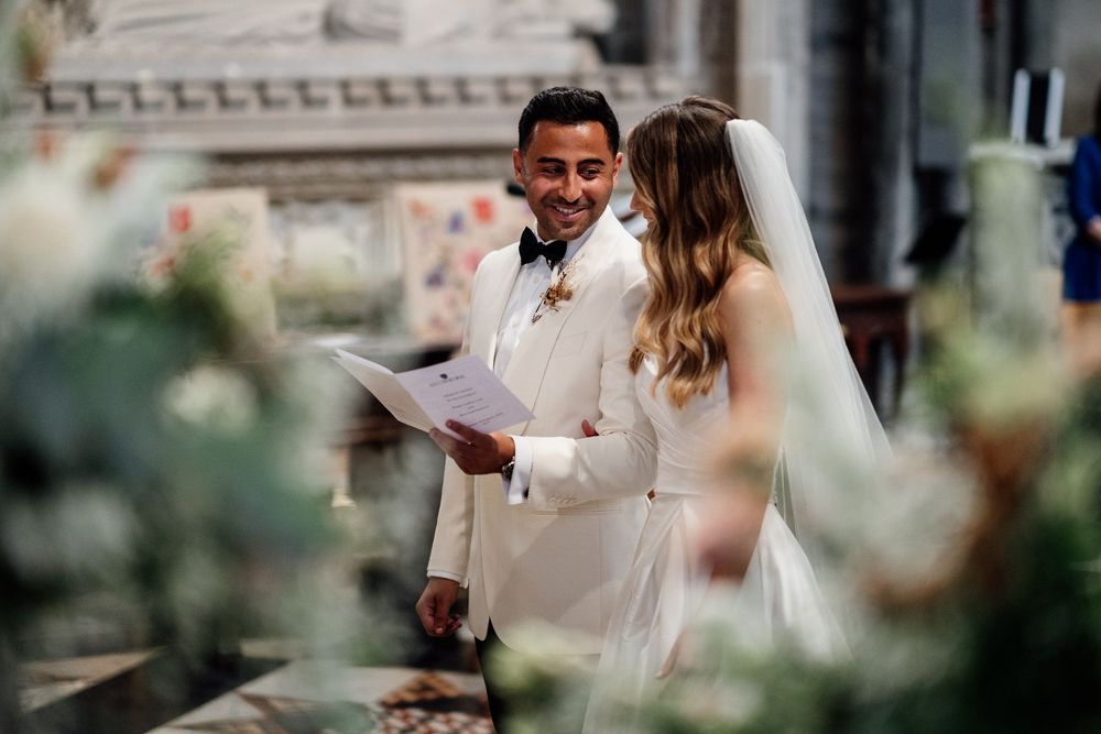 A couple participates in an outdoor wedding ceremony with the officiant reading from papers beneath floral decor.