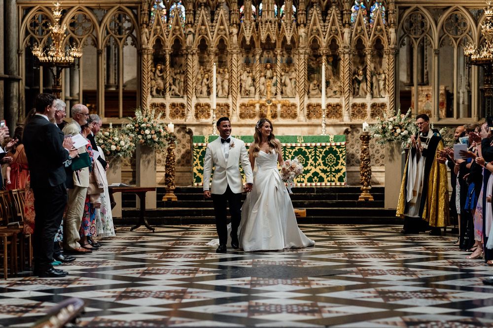 A couple walks down the ornate Ely Cathedral aisle as wedding guests line both sides during an elegant ceremony.