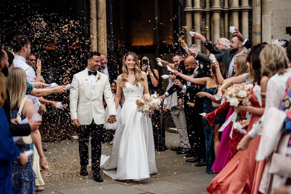 Wedding celebration with guests throwing confetti on a newlywed couple exiting an Ely Cathedral doorway.