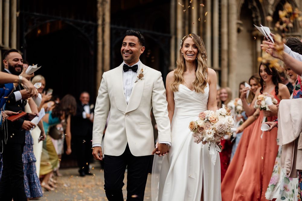 Elegant wedding party celebrates with bridesmaids in coral dresses and groomsmen in black tuxedos at Ely Cathedral.