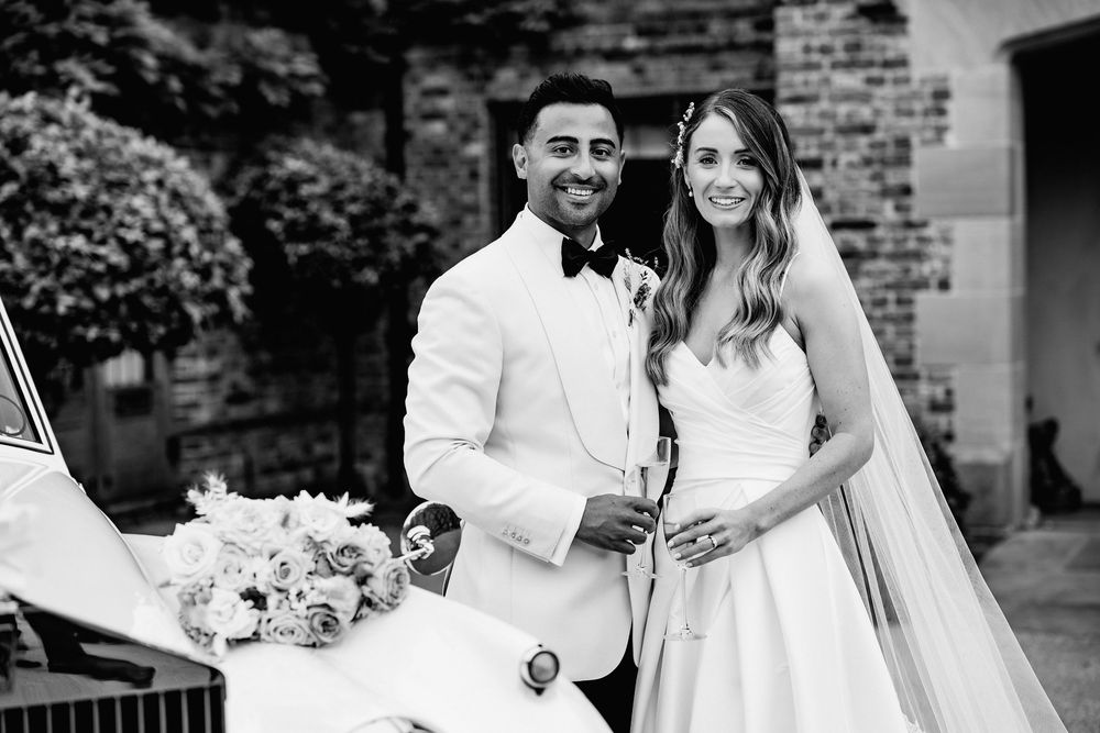 A romantic black and white wedding photo of a couple in formal attire with a bouquet outdoors by a brick wall of The Old Hall.