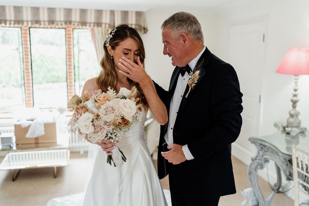 An emotional moment between father and daughter in formal attire, with a beautiful bridal bouquet of blush and white flowers.