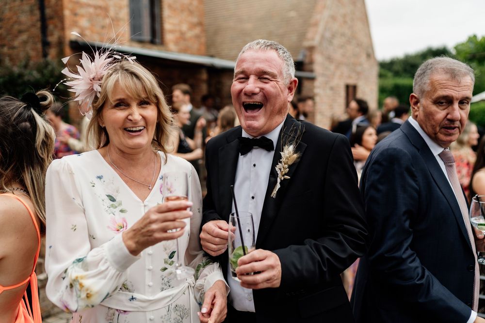 Wedding guests enjoying drinks and laughter at outdoor reception during a joyful celebration in rustic brick setting.