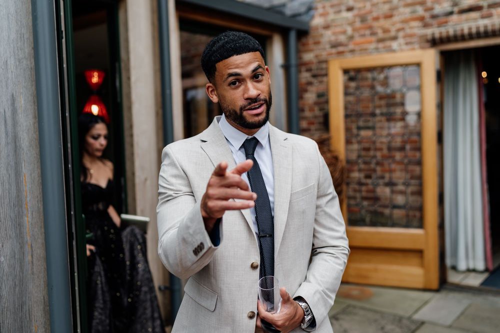 A person in a gray suit jacket gestures expressively while speaking against a brick wall background.