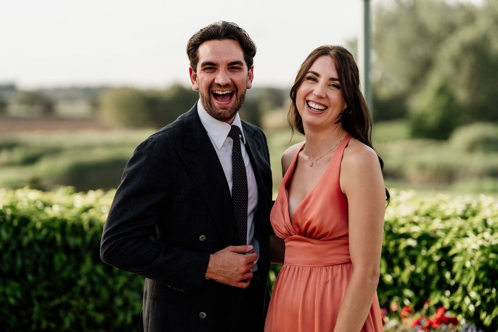A couple in formalwear stands together and smiles at an outdoor event with green foliage in the background.