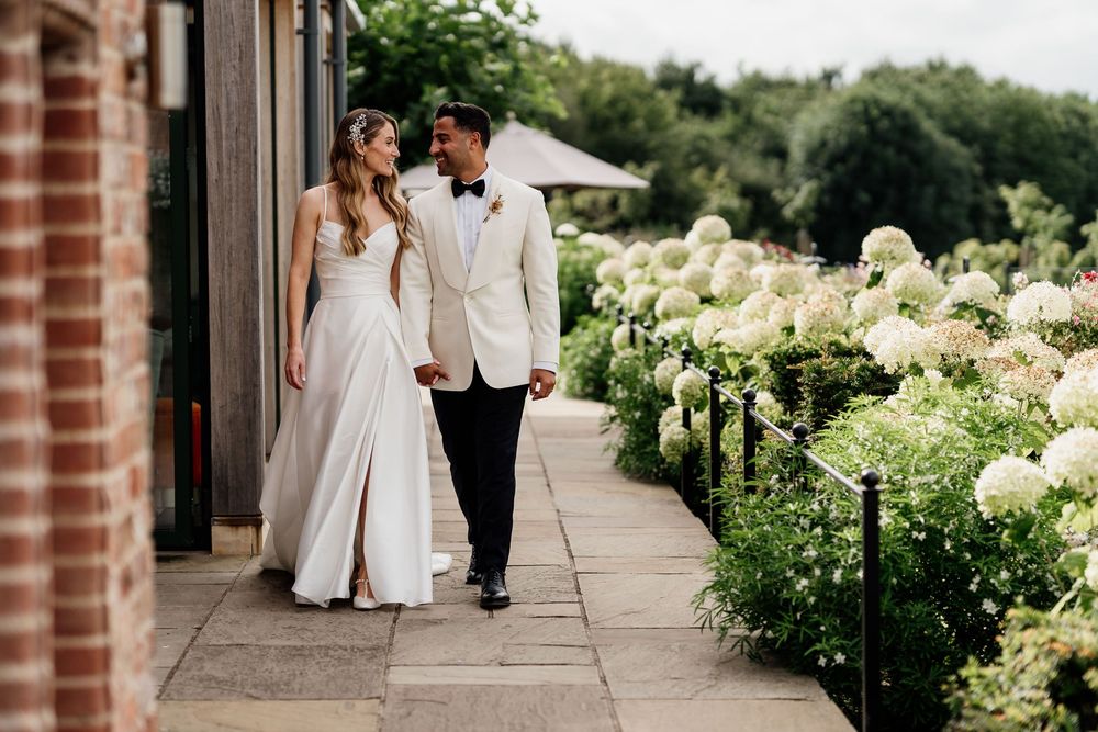A romantic couple walks along a garden at The Old Hall Ely, path lined with blooming white hydrangea bushes and brick columns.