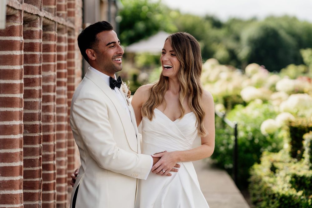 A happy couple shares a joyful moment on their wedding day outdoors by a brick wall with a blooming garden at The Old Hall Ely.