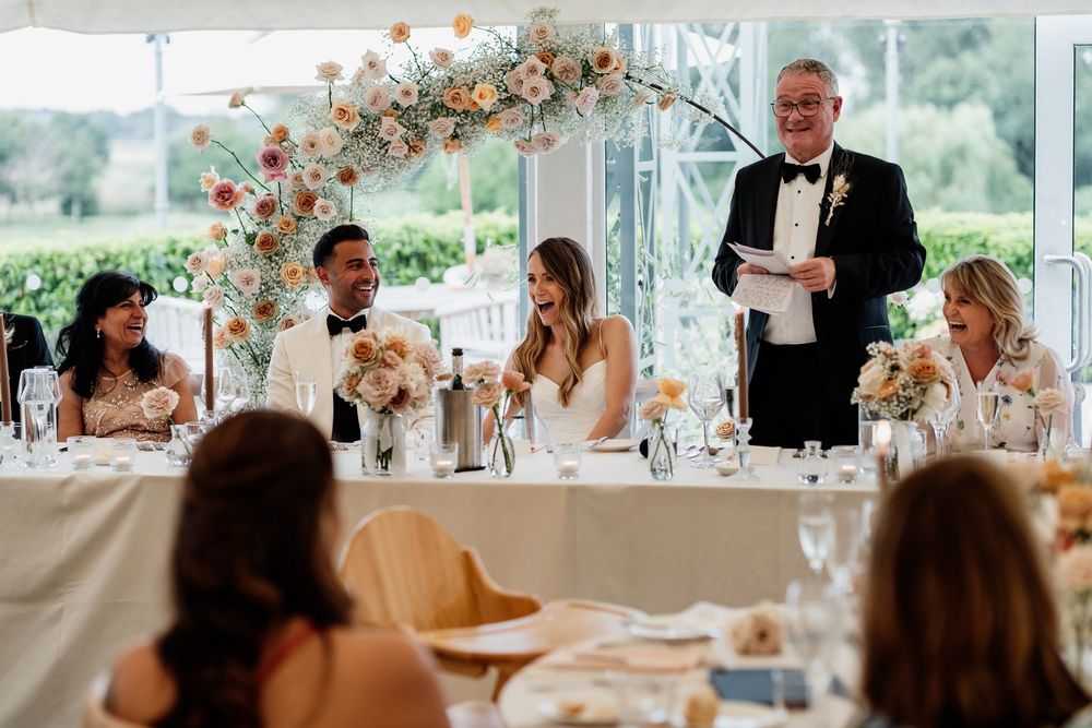 A formal wedding reception with guests seated at decorated tables while someone delivers a speech at the head table.