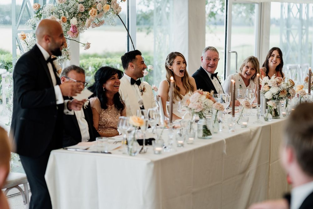 Wedding guests seated at long reception tables in a bright white venue with floral centrepieces and elegant decor at The Old Hall Ely.