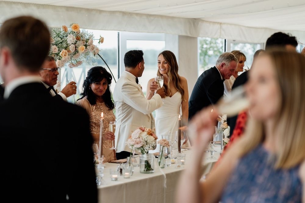 A candid reception scene shows people mingling and conversing at a formal event with white decor and natural lighting.