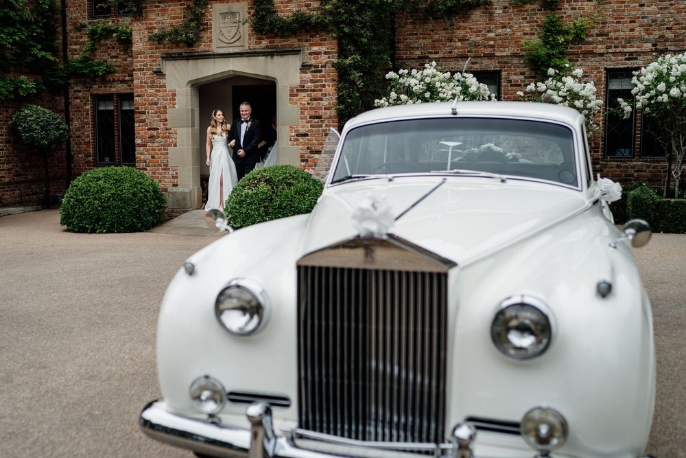 Classic white Rolls Royce parked outside a brick building with decorative shrubs and white floral arrangements.
