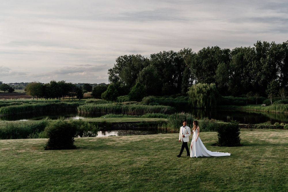 A couple in wedding attire stand together on a manicured lawn with formal garden hedges under a cloudy sky, at The Old Hall, Ely
