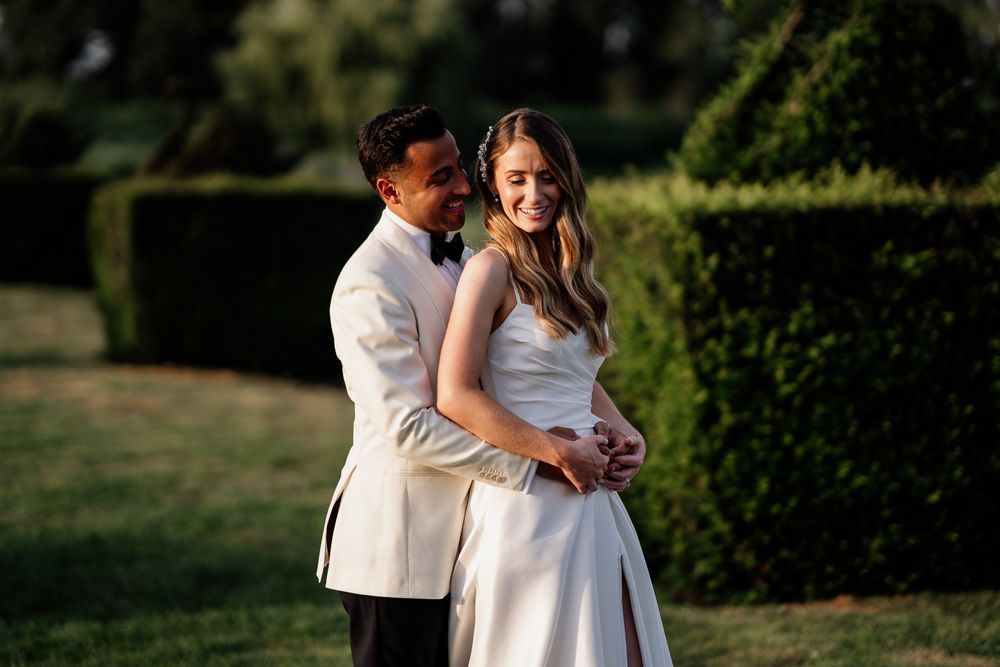 A couple in wedding attire embraces during golden hour in a garden with manicured hedges.