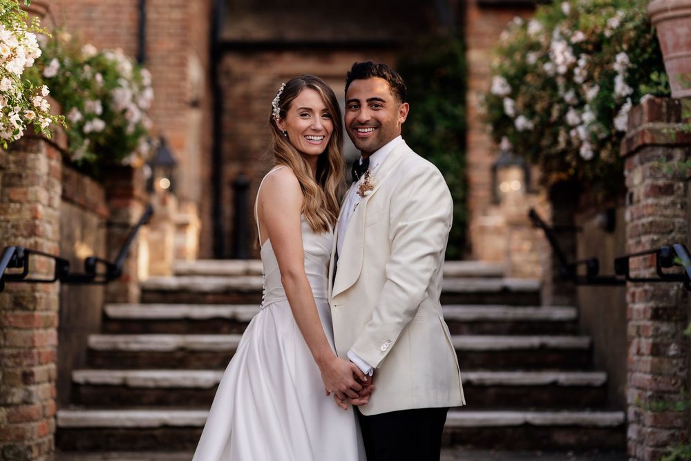 A romantic couple in formal wedding attire poses together at a brick-walled venue with greenery decorations of The Old Hall Ely.