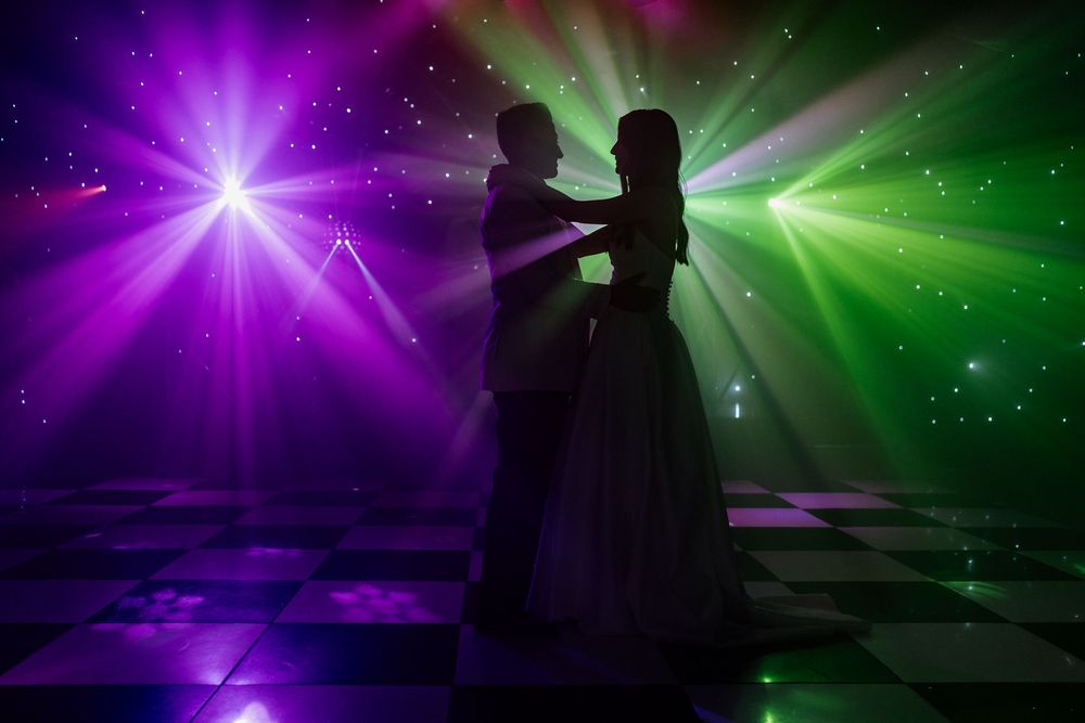 A silhouetted couple dancing under vibrant purple and green lights during a romantic wedding celebration.