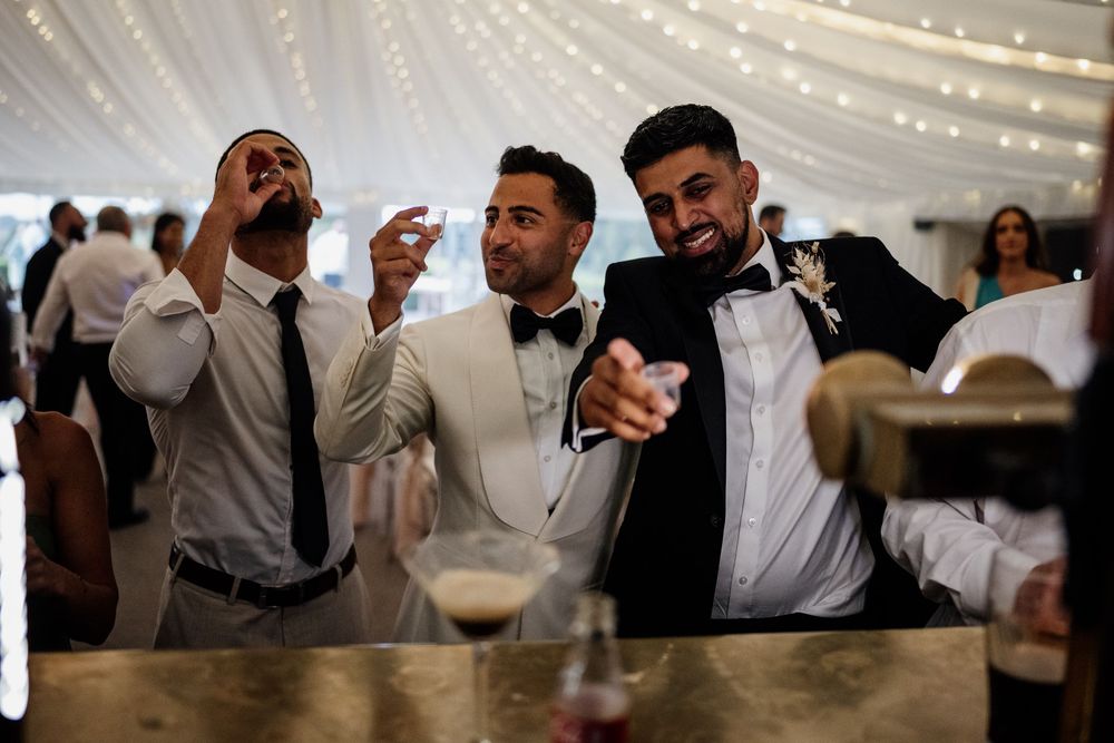 Three smiling guests in formalwear enjoying drinks together at a festive wedding reception under twinkling lights.