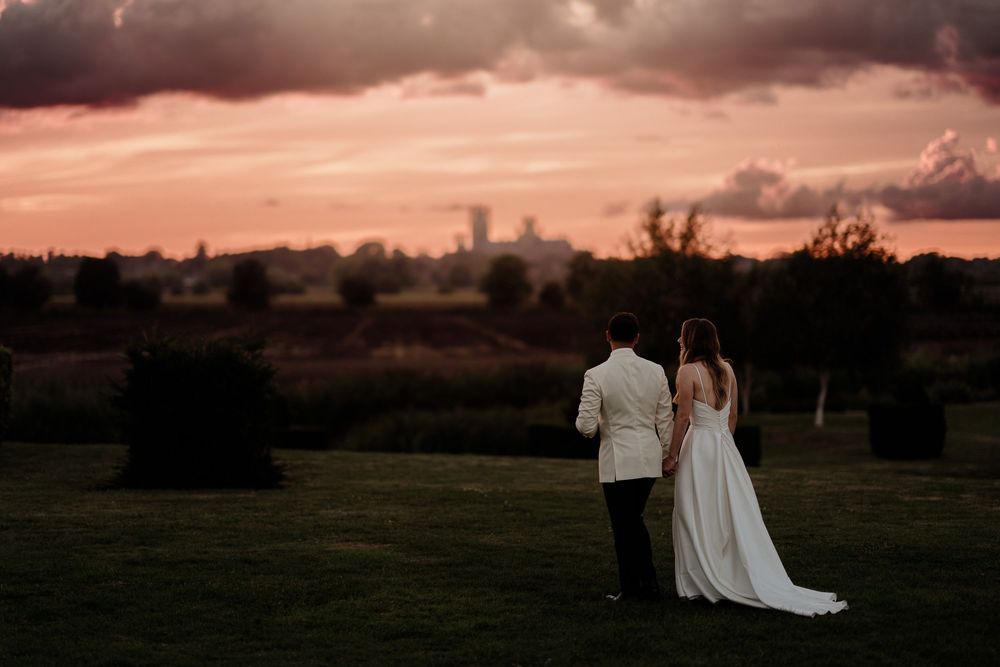 Wedidng Photo with sunset with dramatic clouds cast in golden orange light at The Old Hall Ely, in the distance Ely Cathedral.