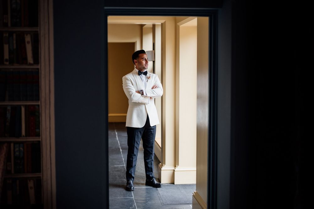Professional in a white blazer and dark pants stands contemplatively in a doorway of an elegant interior.