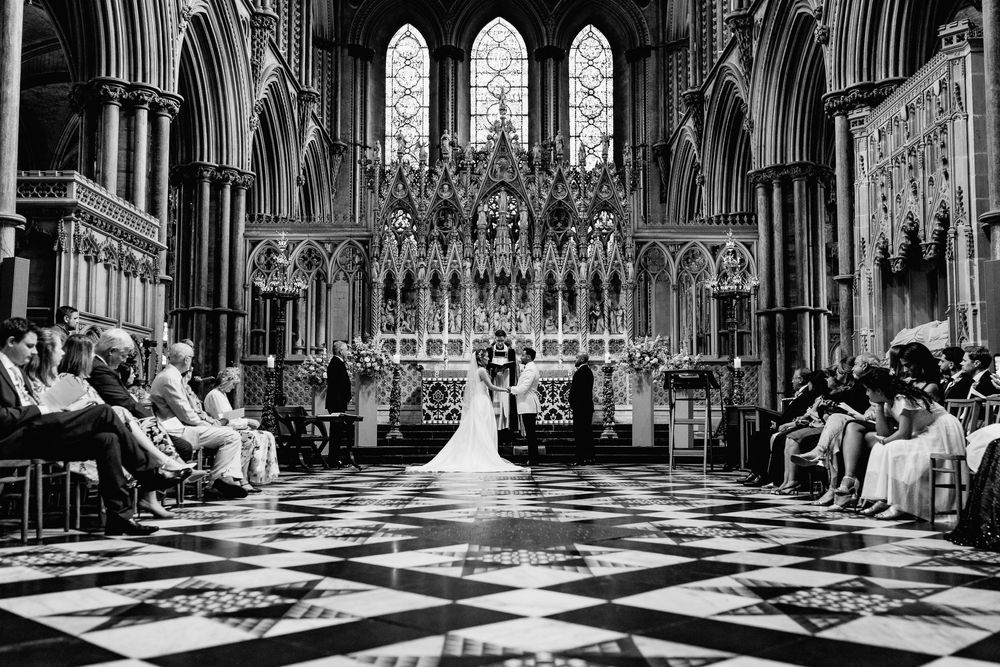 A wedding ceremony takes place in Ely Cathedral with vaulted arches and checkerboard floor tiles.