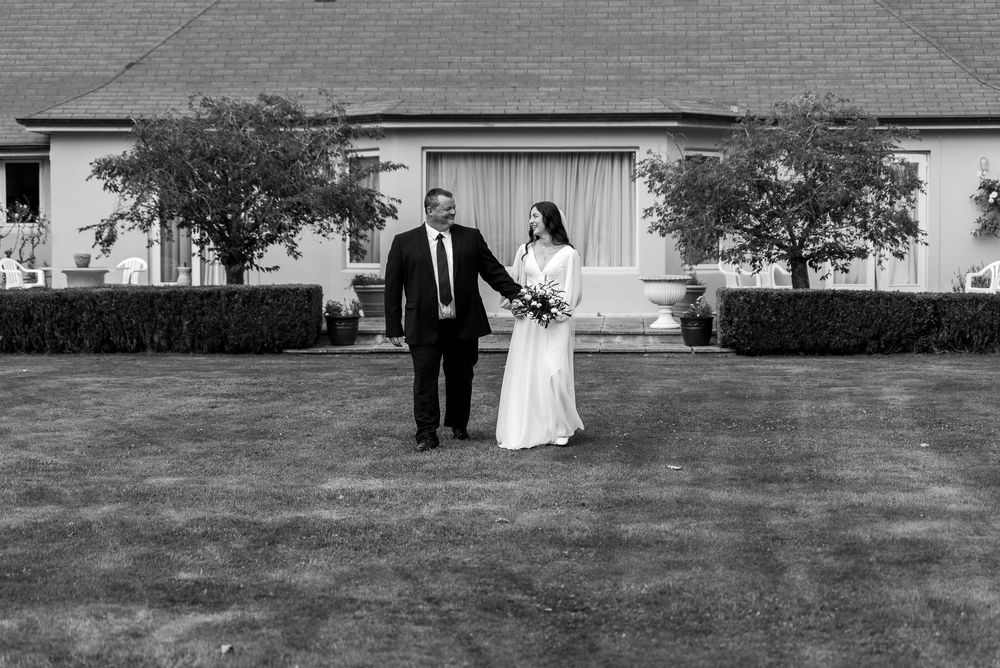 A black and white wedding photo showing a couple walking on a lawn in front of a building with hedges and trees.