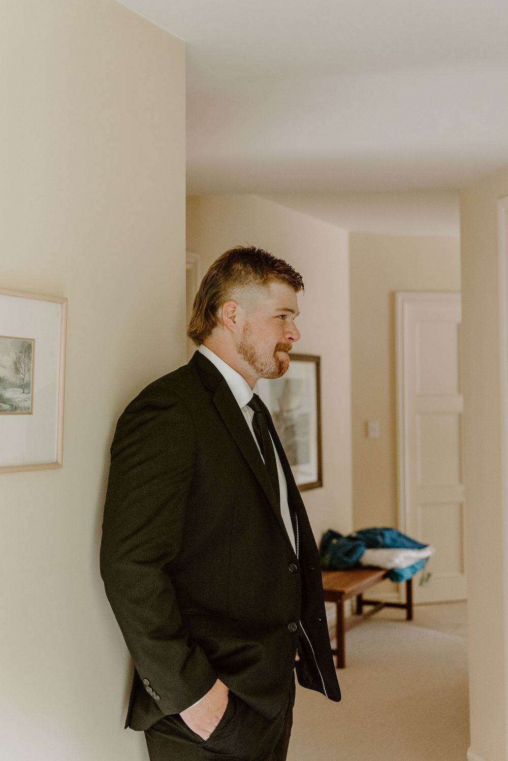 A person in a black suit and tie getting ready in an indoor space with white walls and framed artwork.
