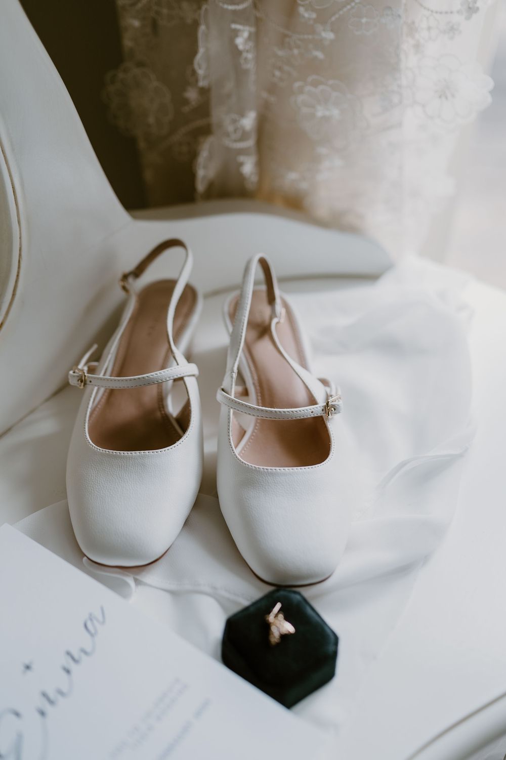White bridal mary jane flats with ankle straps displayed on white fabric next to a small black box.