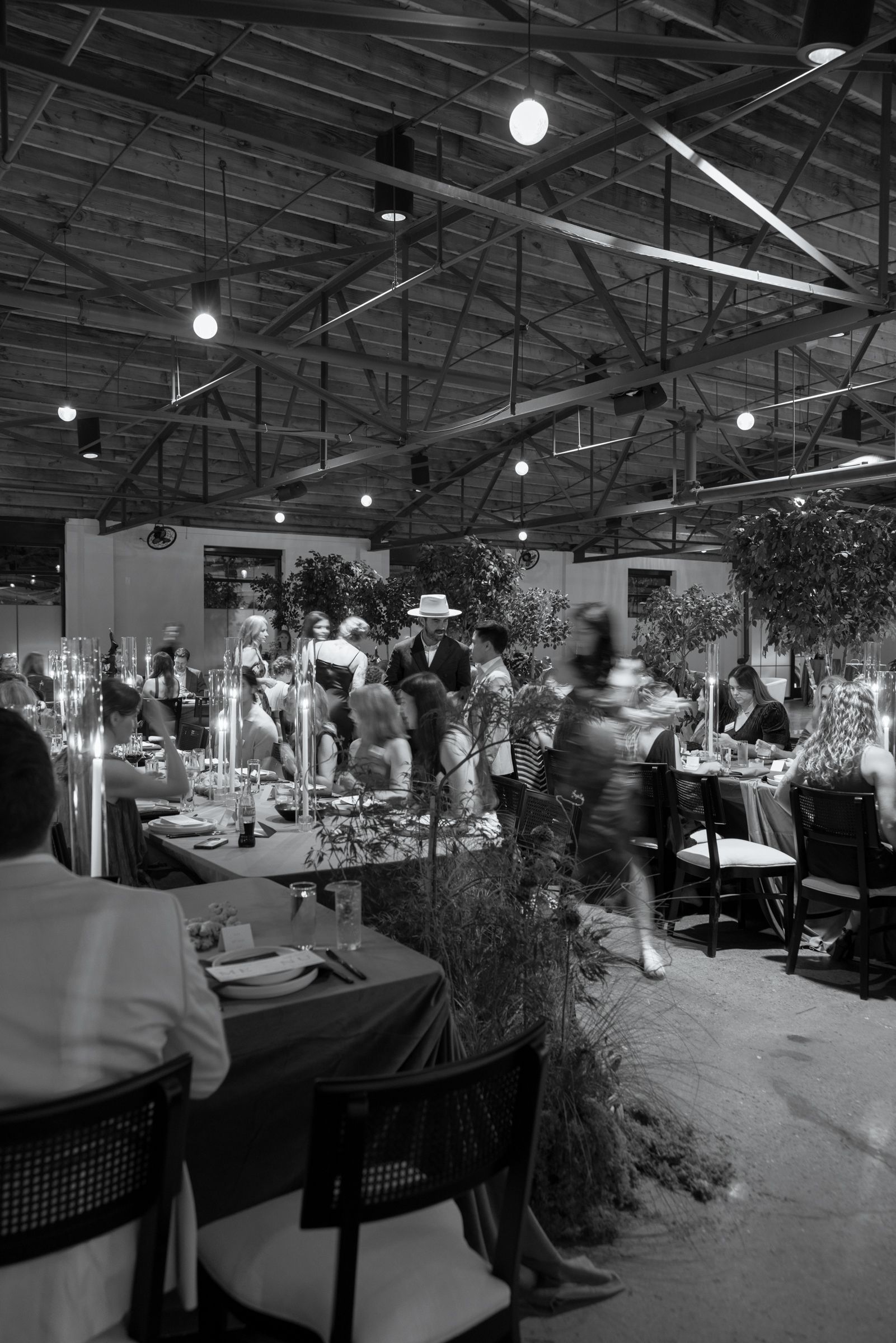 Elegant restaurant interior with high industrial ceiling, hanging lights, and guests dining among potted plants and table settings.