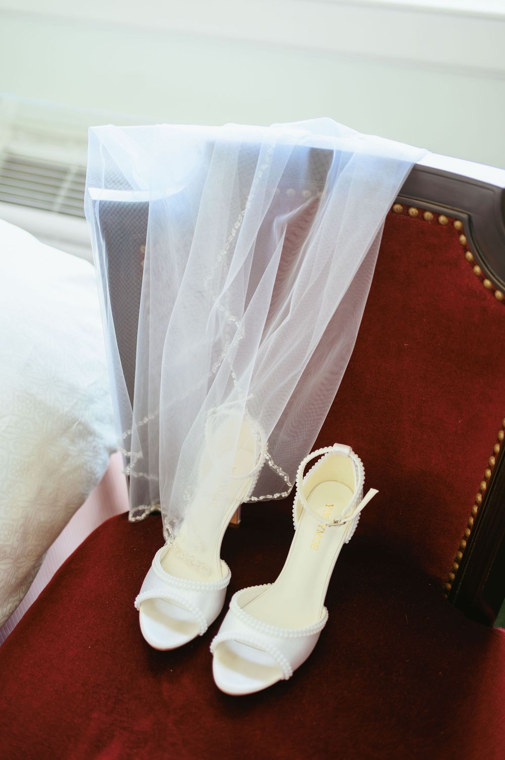 Close-up view of white bridal shoes and tulle veil fabric against a dark red velvet chair.