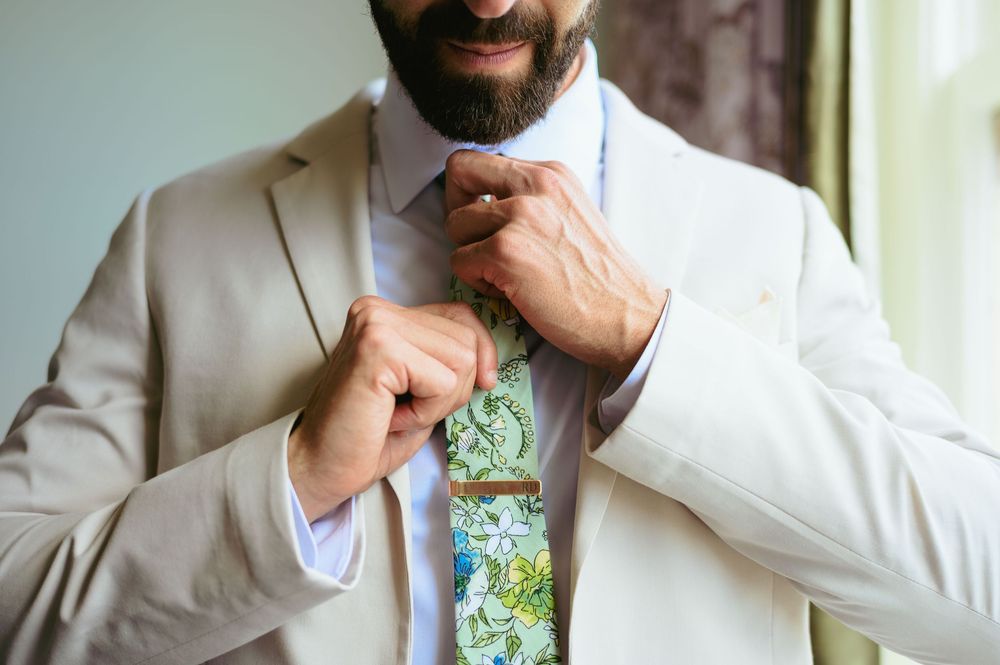 Close-up view of hands adjusting a green floral tie with a crisp white shirt and beige suit jacket.