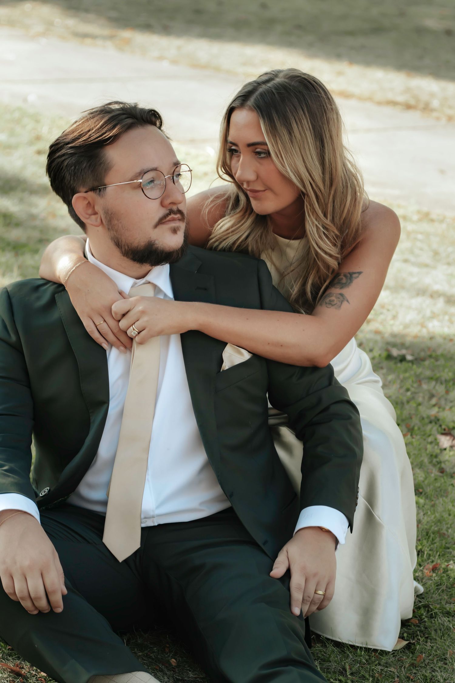 A couple embraces outdoors, with one wearing a forest green suit and beige tie, sharing an intimate moment on the grass.