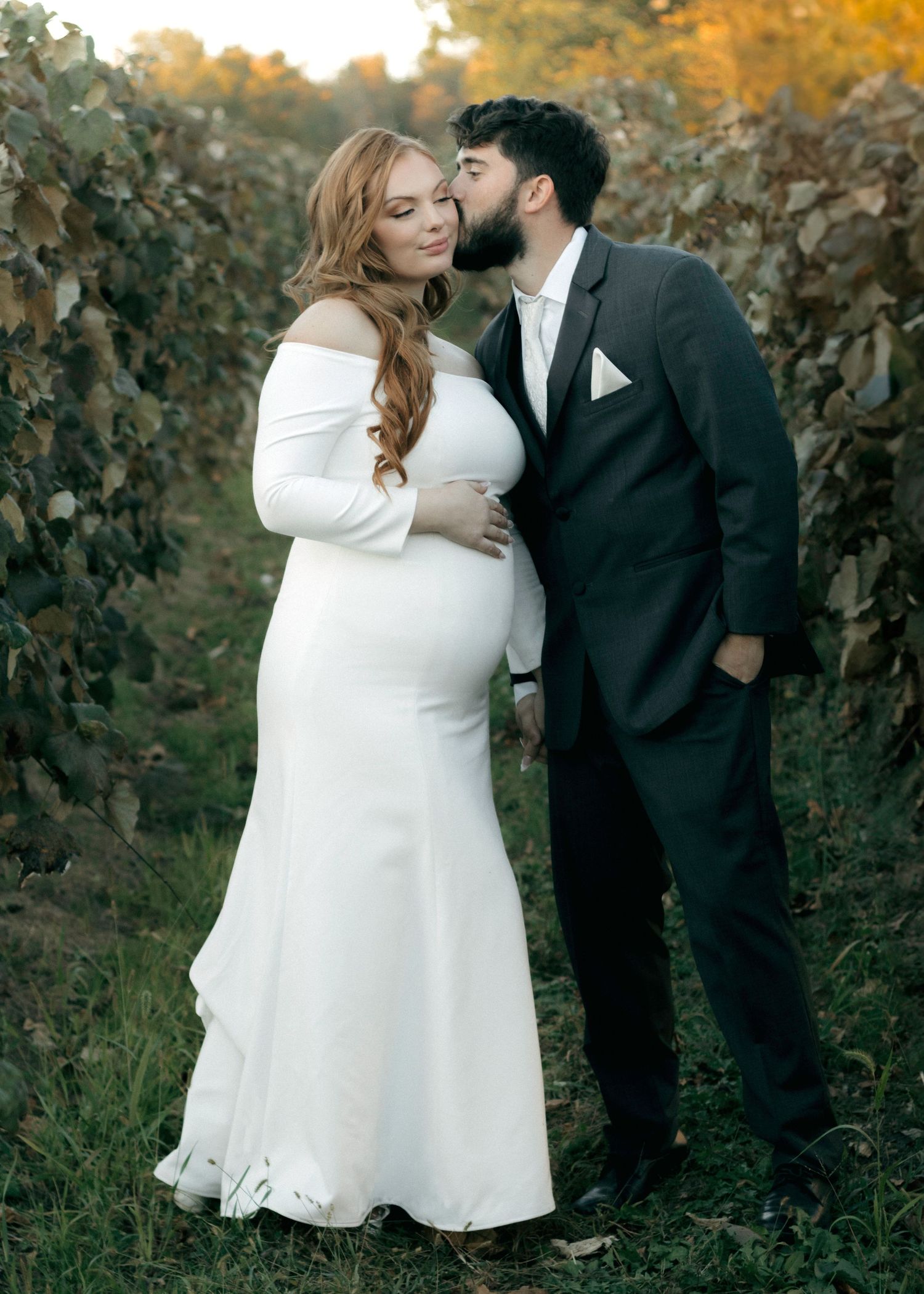 A couple shares an intimate moment among grapevines at sunset, with the bride wearing a long-sleeved white gown.